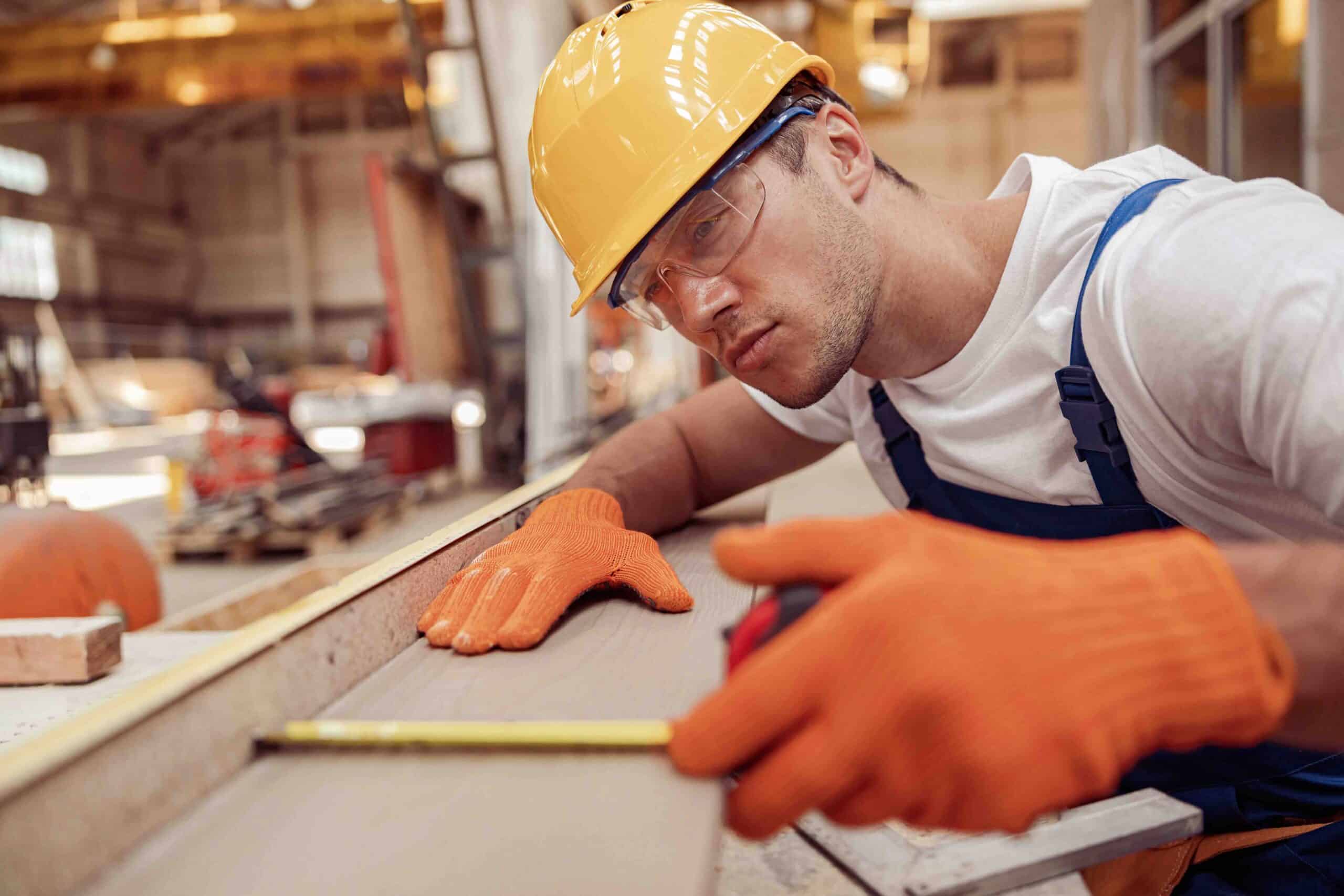 Handsome young man construction worker wearing safety helmet and glasses while placing tape measure on timber plank