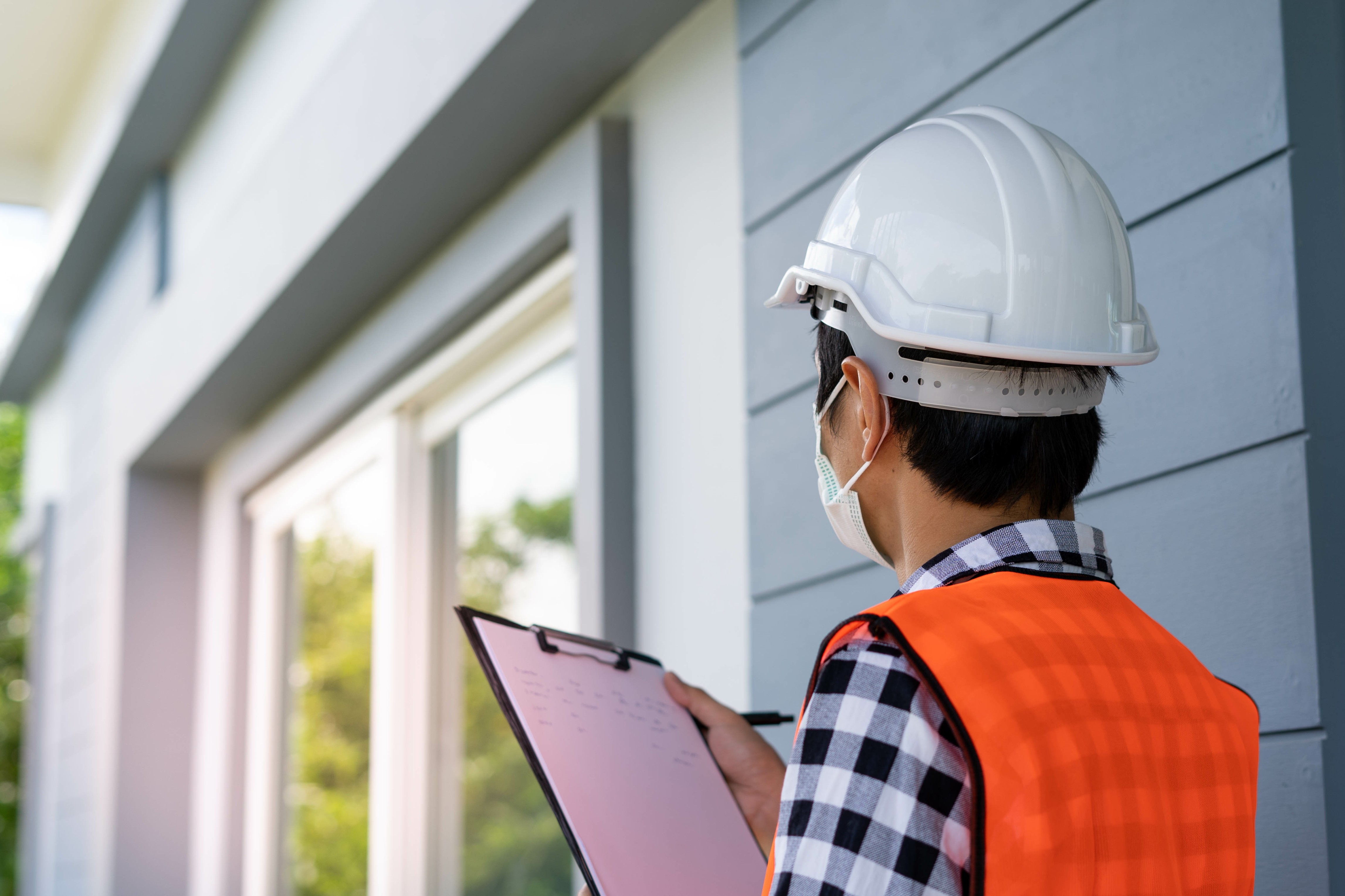 man in a construction hat looking at a house siding and window wall with inspection checklist pad