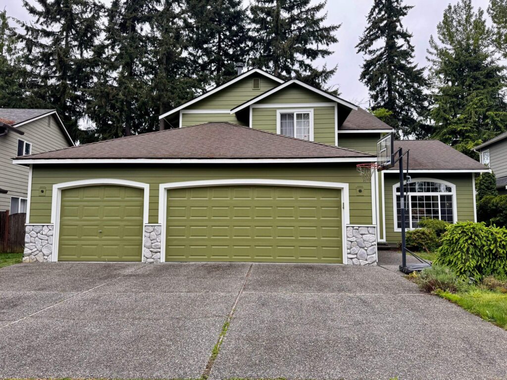 Front view of a two-story suburban home featuring new green James Hardie® lap siding with white trim. The three-car garage has matching green doors framed with white trim and decorative stone accents at the base. A large arched window is visible on the right side, and a basketball hoop stands near the driveway. The house is surrounded by mature evergreen trees and landscaped greenery under an overcast sky