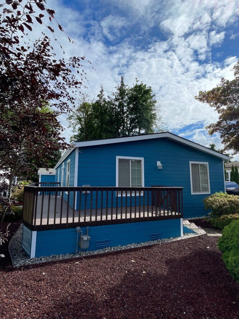 A recently renovated single-story house with vibrant blue James Hardie lap siding, accented by crisp white trim around the windows, corners, and fascia. The home features a dark brown deck railing and is surrounded by clean landscaping with reddish-brown mulch and decorative stones. The bright blue sky and scattered clouds complete the fresh, modern look.