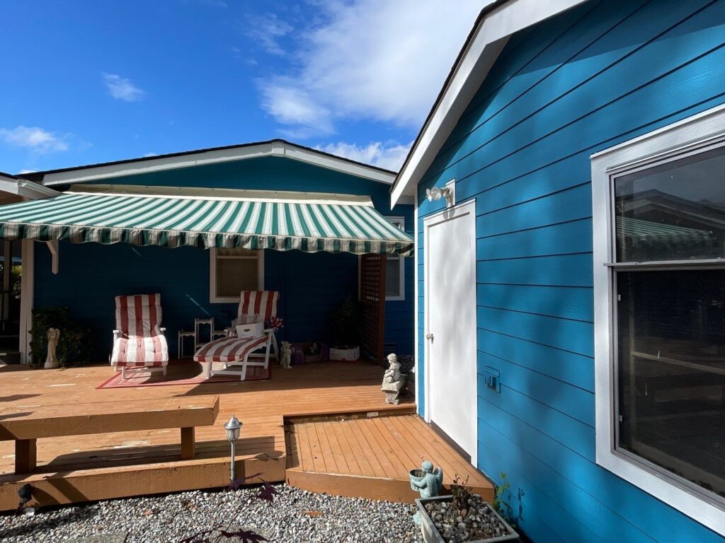 A renovated backyard with vibrant blue James Hardie lap siding and white trim. The outdoor space features a wooden deck with two red-and-white striped lounge chairs under a green-and-white striped retractable awning. Decorative plants, garden ornaments, and gravel landscaping enhance the cozy, relaxing atmosphere. A bright blue sky with scattered clouds adds to the inviting setting.